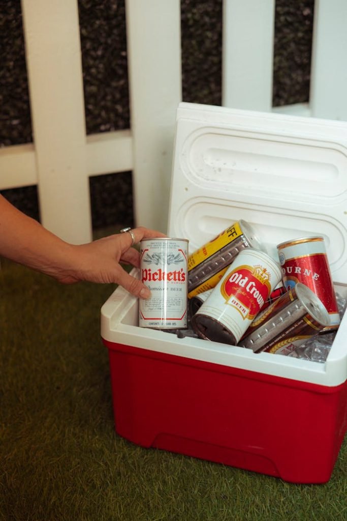 hand-reaching-for-beer-can-in-outdoor-cooler-31208649 A persons hand reaches into a cooler filled with various beer cans outdoors.
